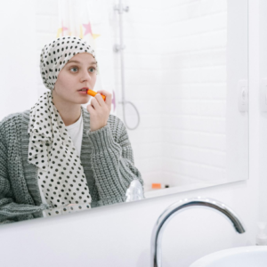 A woman wearing a headscarf applies lipstick while looking in the bathroom mirror.